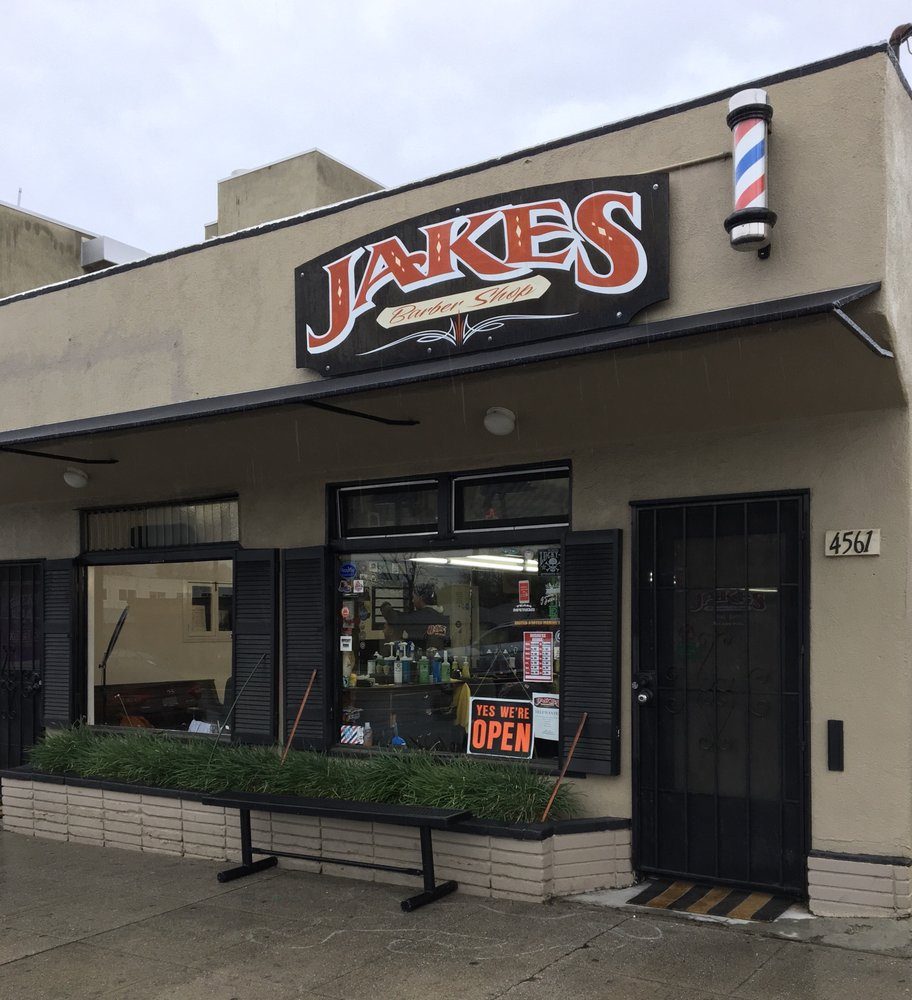 Jake's Barber Shop from the sidewalk: a black sign panel with red JAKES lettering above the window, a striped barber pole at the corner, a bench and a planter under the glass, and a YES WE'RE OPEN card in the window.