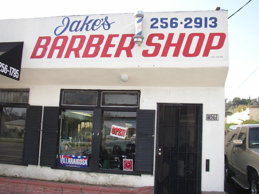 The shop's older painted storefront sign: blue script reading Jake's above red block letters reading BARBER SHOP, with the phone number 256-2913 painted at the right.